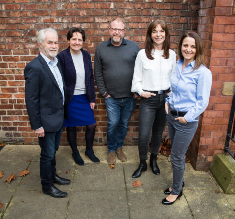 Photo showing 5 people standing in a semi circle smiling in front of brick wall (The CareLoop Team)