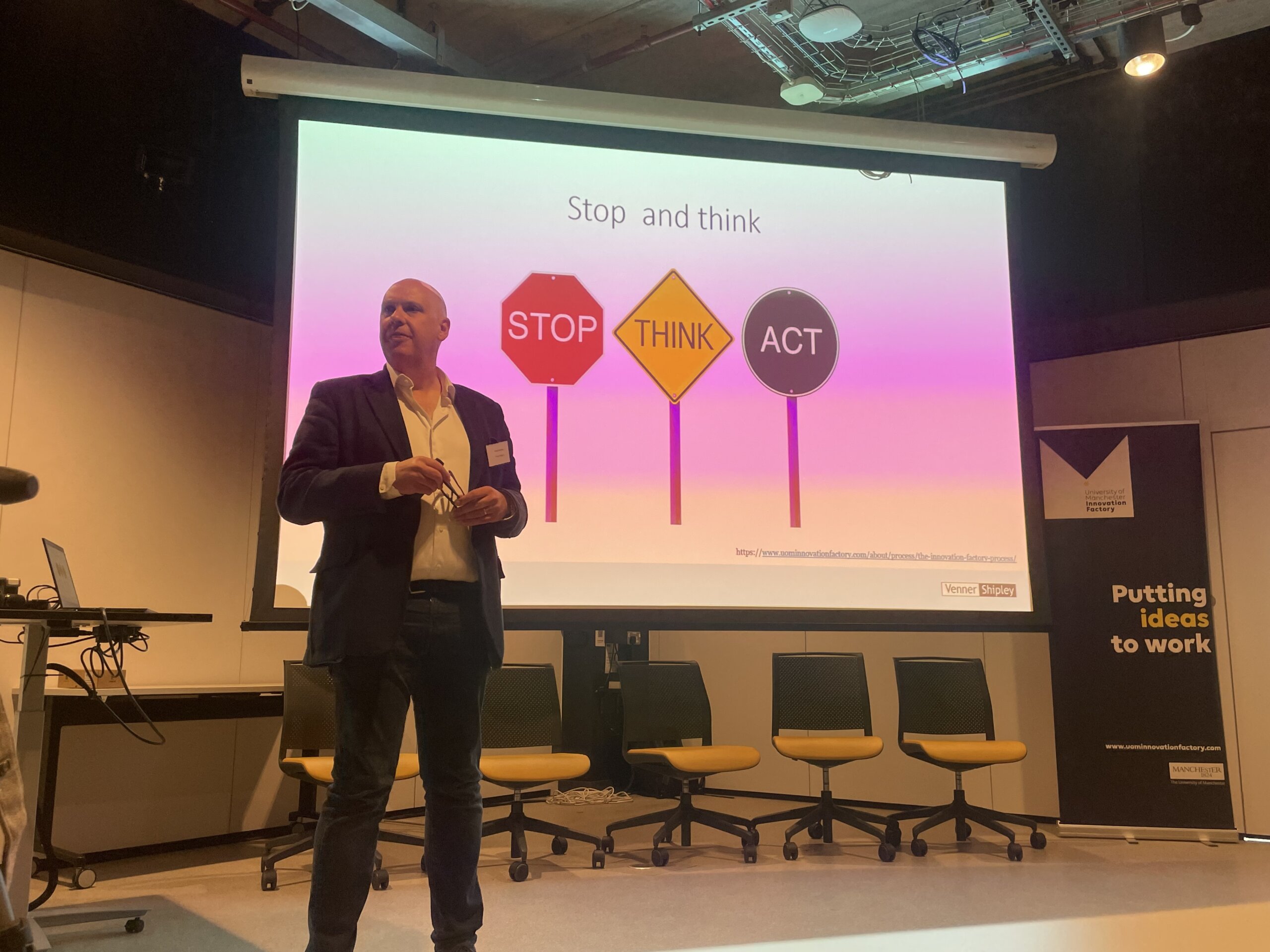 man stands in front of presentation screen which has three signs on it reading Stop, Think, Act 