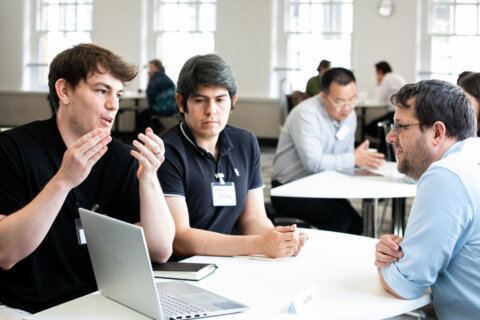3 people sitting at a round table with a laptop talking to each other. in the background there are similar scenes out of focus