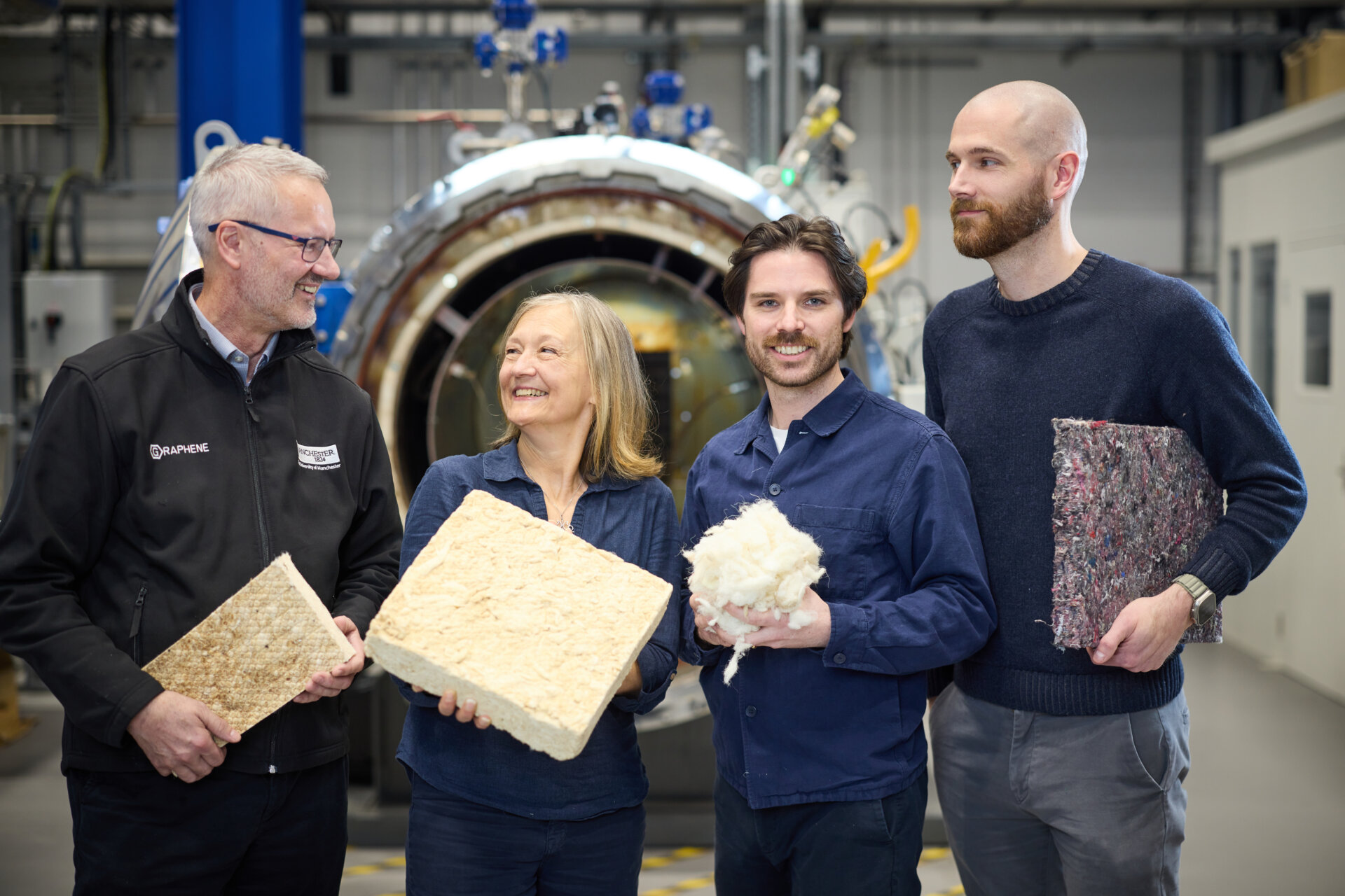 4 people stand smiling at each other and one at the camera. they are in a laboratory wearing blue clothes and golding wool insulation