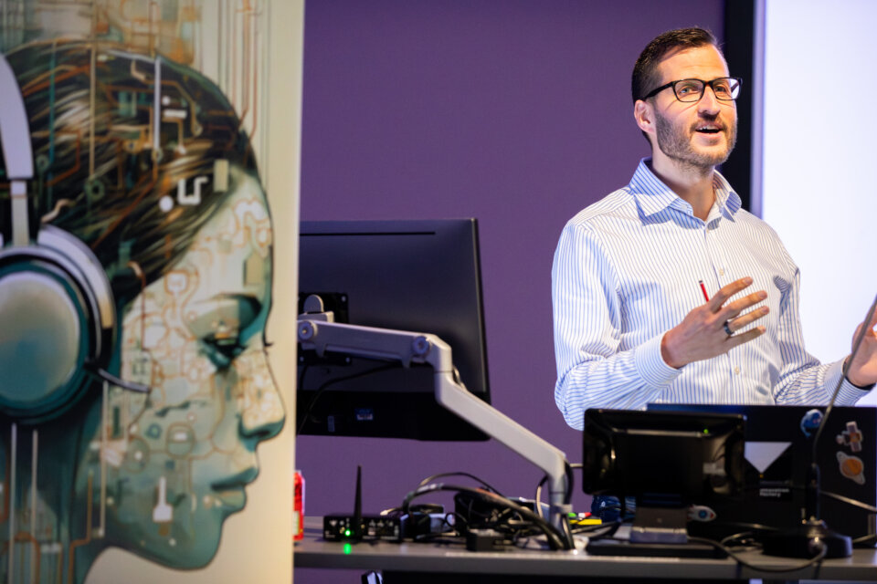 A smartly dressed man with brown hair and glasses presents at a lecturn, in the foreground there is a banner showing the event image - an illustration of a woman wearing headphones