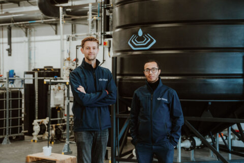 two men stand in front of large tank in a warehouse, looking at camera